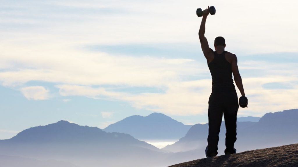 A Man Is Standing At The Mountain With Dumbbells Shows The victory